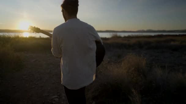 男がギターを持って湖に向かって歩いて絵のように美しい夕日を見る 横4Kビデオ — ストック動画