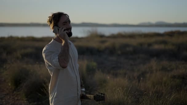 携帯電話で通話するクールな男の肖像画を閉じ 湖の近くの日没時に手にアコースティックギターを持っているうなずいて 水平方向の4Kビデオ — ストック動画
