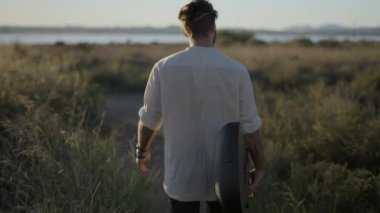 Hipster bir adamın elinde gitarla gün batımında çimenli bir arazide yürümesi - ardından - 4K yatay görüntü