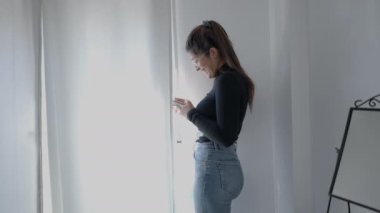 Kolombiya Üniversitesi 'nin genç bir öğrencisi çalışmaya ara verdi. Pencerenin önünde duran ve telefonunu kullanan genç bir kız. - 4K Yatay Görüntü