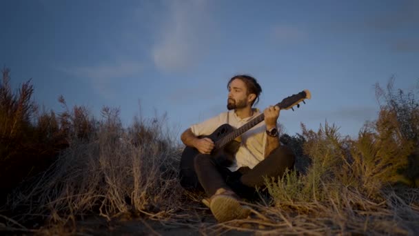 芝生の牧草地に座り 日没時にギターを弾く若い男 スローモーションプルバック 横4Kビデオ — ストック動画