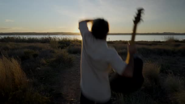 湖の上の夕日を見るギターでジャンプする男のバックビュー 水平4Kビデオ — ストック動画