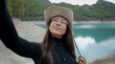 Şapkalı ve gözlüklü genç bir kadının gölün kenarında selfie çekişini yakından izleyin.