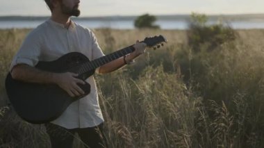 İspanyol gitarist sonbaharda çayırlarda akor çalıp Torrevieja Pembe Gölü yakınlarındaki siyah akustik gitarda şarkı söylüyor - Yatay 4K video