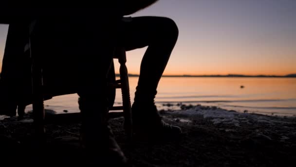 日落时在湖边用吉他弹奏和弦的嬉皮士的轮廓 男子吉他手在暮色的时候坐在椅子上打腿和即兴弹奏的时候 水平4K视频 — 图库视频影像