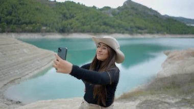 Koyu renk saçlı genç kadın gölün yanında selfie çekiyor, içeri itiyor ve dışarı itiyor.