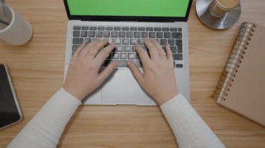 Ofis masasının üstünde yeşil ekranlı dizüstü bilgisayarı ve klavyeye yazı yazan kadın elleri var. Gözlük, kalem ve cep telefonu yanında. Krom anahtar. - Yatay 4K videosu