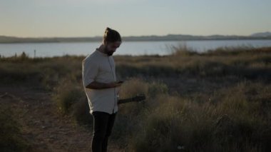 Gitarist bir gitarist gün batımında nehir kenarında dikilirken cep telefonuyla arama yapıyor.