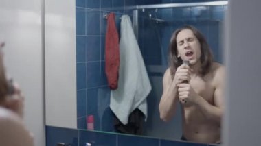 Uzun saçlı bir adam, mikrofon olarak saç fırçasıyla banyo aynasının önünde şarkı söylüyor.