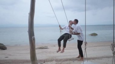 Lovely male gay couple honeymooners kissing while riding a beach swing at tropical island at dusk - 4K Horizontal video