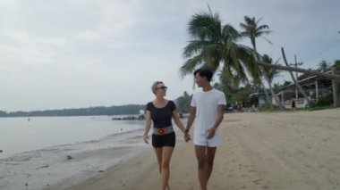 Irklar arası ve kültürler arası güzel bir çift el ele yürüyorlar birbirlerine bakıyorlar ve Tayland 'da egzotik bir adanın sahilinde konuşuyorlar - 4K Yatay Video