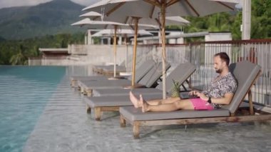 Orta yaşlı yetişkin bir adam dizüstü bilgisayarında gezerken egzotik bir tatilde sonsuzluk havuzu kenarındaki bir iskemlede yatıyordu. Görüntü - profil - 4K Yatay Görüntü