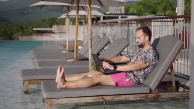 Orta yaşlı, sakallı bir adam dizüstü bilgisayarlarda gezinirken tropikal tatil beldesinin yanındaki havuzun kenarındaki koltuğa uzanmış. Tatil - 4K Yatay Görüntü