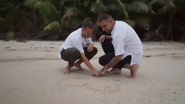 Happy Gay Couple Honeymooners Squatting Position Draw Heart Shape Sand — Vídeo de Stock