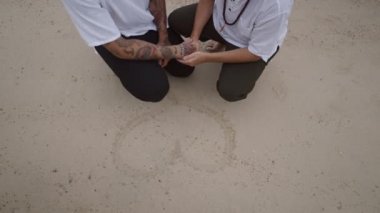 Male Gay Couple on the beach in the squatting position holding hands by the heart shape handwritten on the sand - 4K Horizontal video