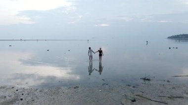 Irklar arası gezginler, gün batımında ada sahilinde sığ deniz suyunda el ele yürüyorlar. Havada dönen 4K yatay video.