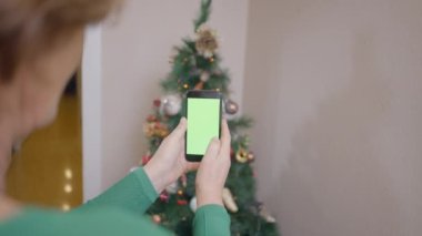 Yeşil ekranlı cep telefonu tutan yaşlı kadın, evin köşesinde Noel ağacının fotoğraflarını çekiyor. - Ekin görüntüsü - 4K yatay görüntü