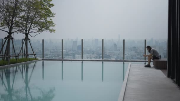 Lone Guy Using Smartphone Sitting Poolside Lounge Cityscape Background Static — Stok video