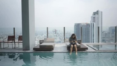 Millennial Nomad, Tayland 'daki Bangkok Metropolis' in panoramik manzarasına bakan otelin yakınındaki bir videoyu çekerken şezlongda oturuyor. Statik - 4K Yatay Görüntü