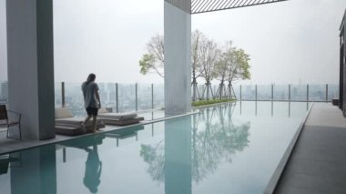 Adam, akıllı telefonuyla konuşurken havuz kenarındaki güneşli yatakta oturuyor. Geniş Görüntü - 4K Yatay Görüntü