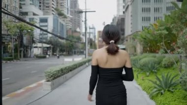 Kısa siyah elbise giyen ve maske takan genç, güzel bir Asyalı kadın boş bir iş caddesinde neşeyle yürüyor. - 4K yatay video