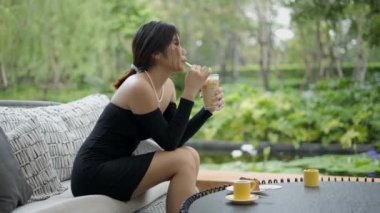 Güzel, Asyalı bir iş kadınının, elinde bir fincan kahveyle güzel yeşil bir arka planı olan bir otelin lobisinde seksi, siyah bir elbise giydiği bir film çekimi. - 4K yatay video