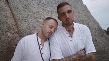 Gay couple with closed eyes resting leaning on each other shoulder by the rock looking at the camera - 4K Horizontal video