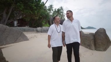 Lovely married gay couple honeymooners walking holding hands along the rocky beach by the sea and talking with a happy smile on a face - 4K Horizontal video