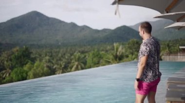 Tatil gezisi sırasında arka planda tropikal palmiye ormanları ve dağları olan sonsuz havuz kenarındaki cep telefonuyla konuşan genç bir yetişkin - 4K Yatay Video