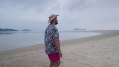 Tayland 'da kasvetli bir günde, tropikal bir adanın kumlu sahilinde yürüyen, yaz kıyafetleri içinde orta yaşlı yetişkin bir adam.
