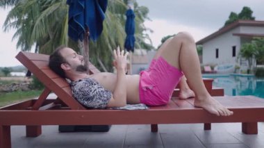 Bearded man traveler watching video on phone while resting on wooden deckchair by the pool in tropical hotel - 4K Horizontal video