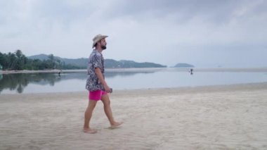 Kasvetli bir günde 30 'lu yaşlarda hippi sakallı bir adam Tayland' da ada sahilinde yürüyor - ağır çekim - 4K yatay video