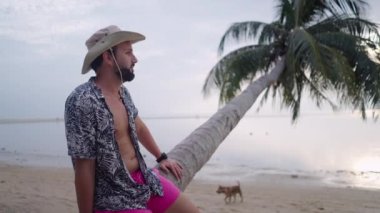 Orta yaşlı sakallı bir gezgin eğilmiş avuçlarında oturuyor ve Tayland sahilinde günbatımının tadını çıkarıyor - 4K Yatay video