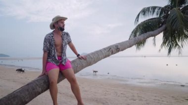 Gün batımında deniz kenarındaki tropik sahilde eğilmiş palmiye üzerinde dinlenen beyaz bir adam. 4K Yatay video.