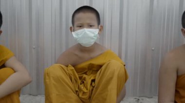 Bangkok 'ta metal çitin yanında oturan turuncu cüppeli ve solunum maskeli genç bir çocuk. 4K yatay video.