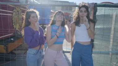Three dark-haired girls with lollipops by metal fence on windy day - 4K Horizontal video