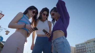 Dark-haired girls in sunglasses stand outdoors and use phones and talk - 4K Horizontal video