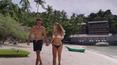 Sahilde yürüyen genç bir çiftin yan görüntüsü. Kız bikinili ve oğlan çıplak vücutlu ve kısa - 4K yatay video