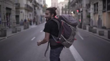 Handsome backpacker running in the middle of the street smiling and having fun, gives a look at the camera and continues running. - Horizonal video