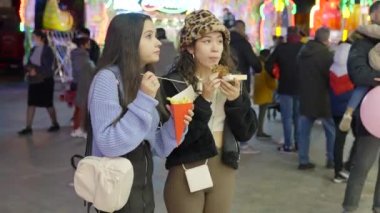 Two young women talking while eating snacks at night Valencia funfair market with many people on background - 4K Horizontal video