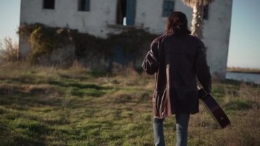 Man flipping his hair and holding guitar walking towards abandoned house in slow motion - Horizontal video