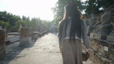 A young teenager walking by a row of concrete Buddha Statues standing on a brick base against bright sun in Wat Yai Chai Mongkhon temple in Ayutthaya, Thailand - back view - 4K Horizontal