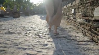 Legs of the woman in dress and slippers walking on stone pavement road against the sun at Wat Yai Chai Mongkhon temple in Ayutthaya, Thailand - 4K Horizontal