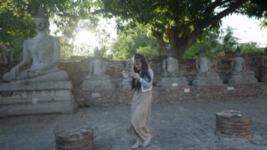 Brunette girl using phone while visiting Wat Yai Chai Mongkhon temple in Ayutthaya at sunset, Thailand - 4K Horizontal
