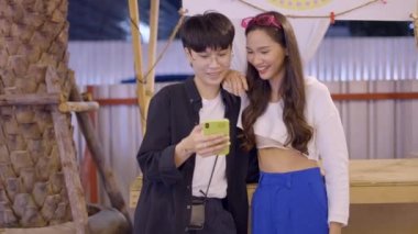 There is a couple standing somewhere. The girl is wearing a black shirt that is open and a white T-shirt inside it surfing mobile and showing it to her partner. - 4K Horizontal video