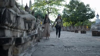 Two Thai girls travel inside Wat Yai Chai Mongkhon temple in Ayutthaya at sunset walking along a row of concrete Buddha statues, Thailand - 4K Horizontal