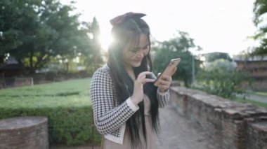 Cute Happy Asian young girl using a smartphone at sunset against setting sun - slow motion - 4K Horizontal
