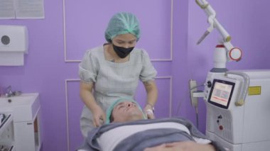 Cosmetologist puts on hygienic cap on patient and applies face cream before cosmetic treatment - 4K Horizontal video