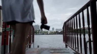 Male Travel Photographer Walks Towards Chao Phraya River With DSLR Camera To Capture Fascinating Cityscape Of Bangkok, Thailand. dolly-out shot - 4K Horizontal video