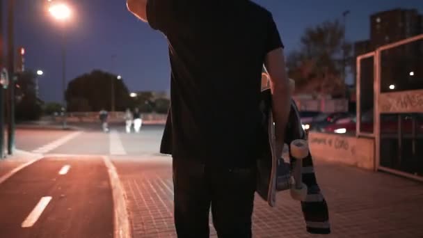 夜にスケートボードを運ぶ横断歩道に向かって歩く若い男 4K水平ビデオ — ストック動画
